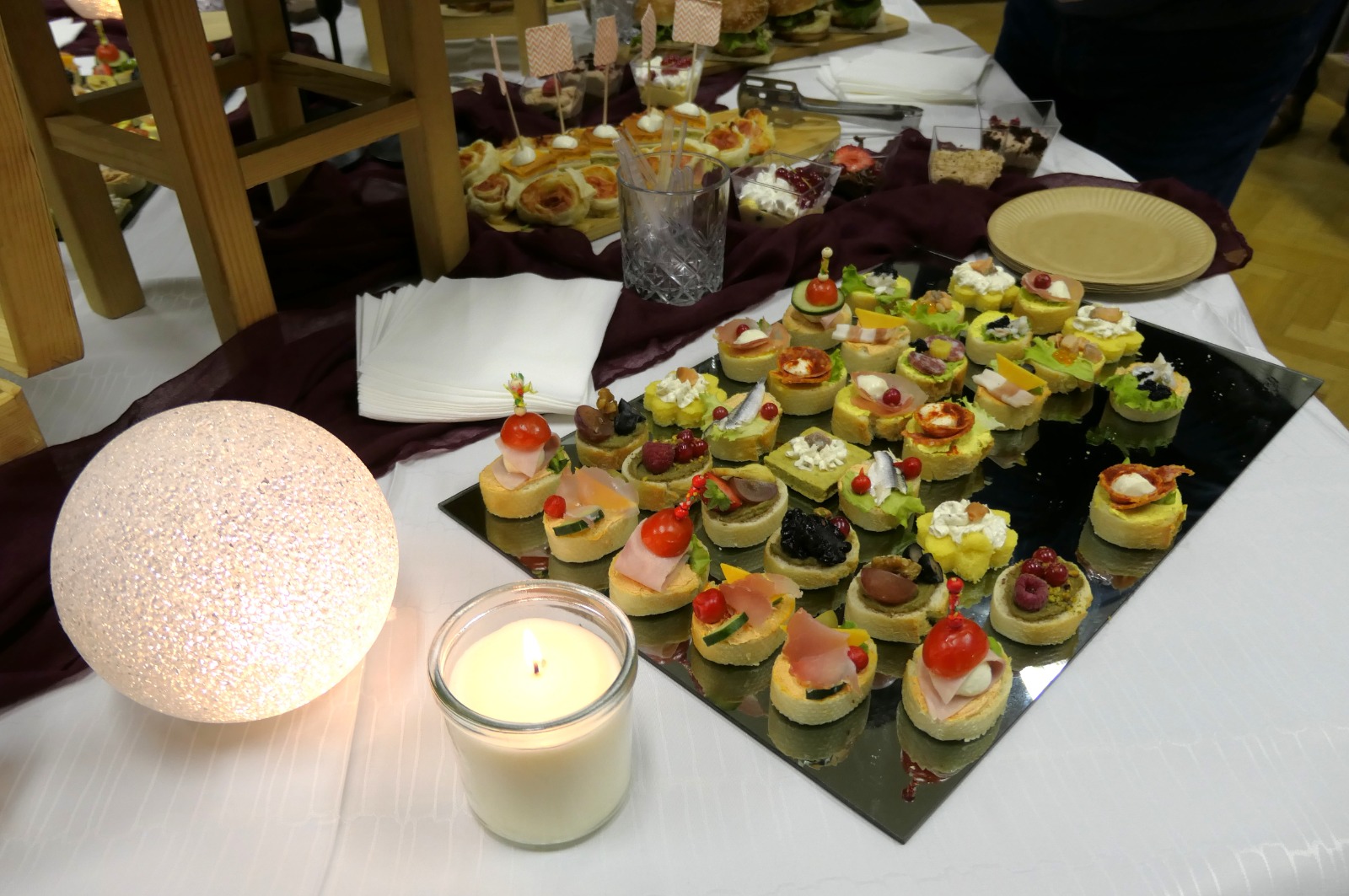 Tavolo con antipasti decorati, tra cui mini tartine con ingredienti vari e decorazioni colorate, accompagnato da una candela e una lampada luminosa.