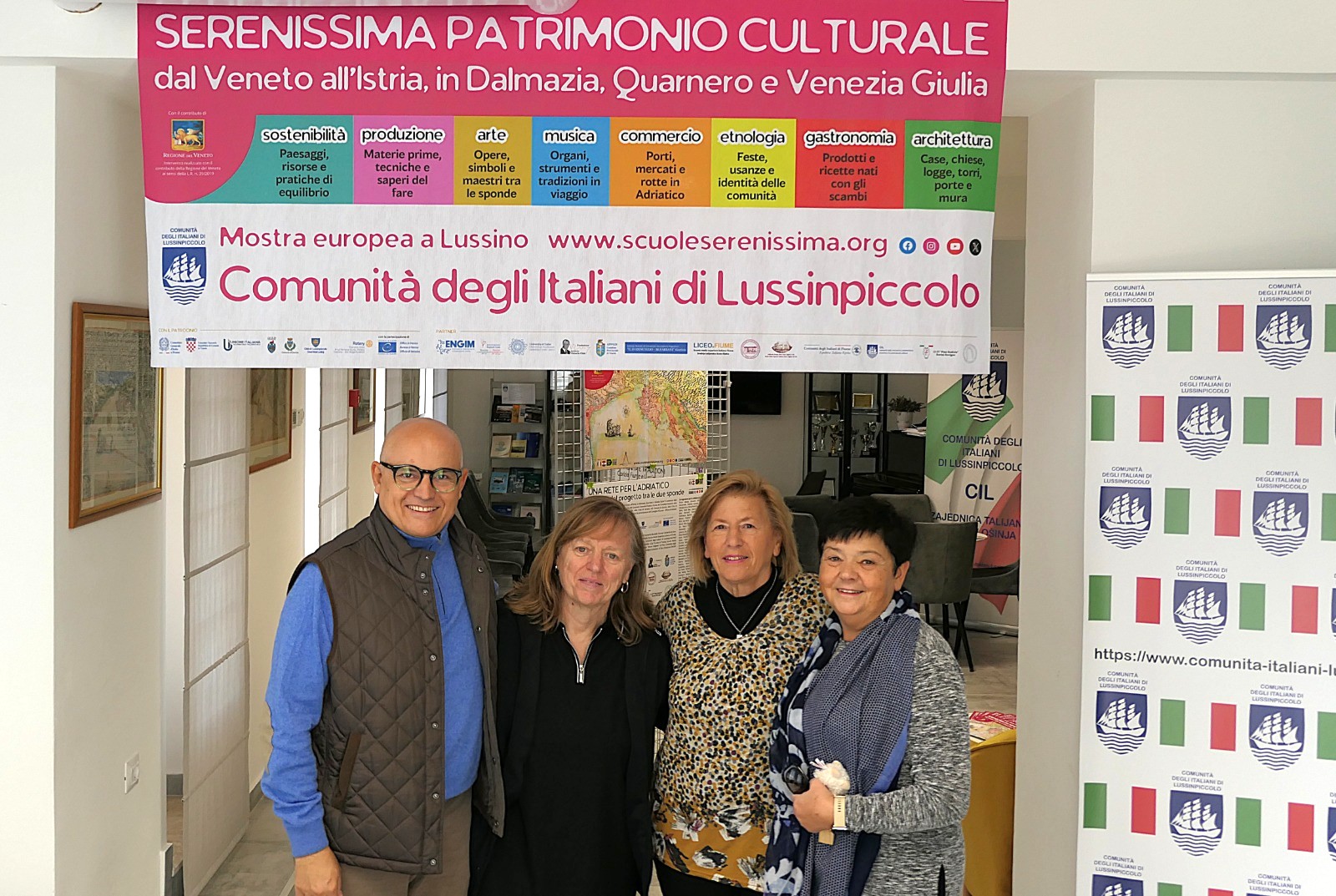 Un gruppo di quattro persone sorridenti che posano davanti a un grande striscione che promuove il patrimonio culturale della Serenissima, in un ambiente riconoscibile come un centro comunitario.