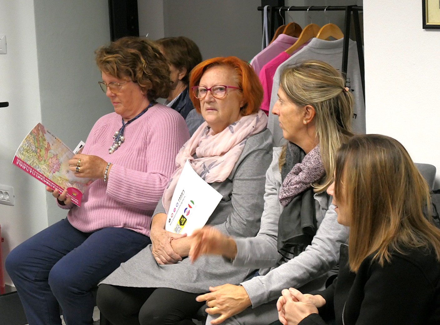 Gruppo di donne sedute in una riunione, alcune leggono documenti e discutono. Lo sfondo mostra alcuni vestiti appesi.