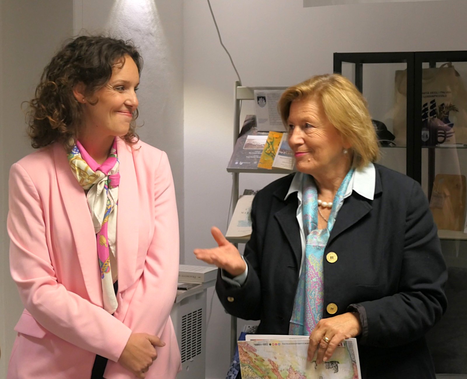 Due donne conversano in un ambiente interno. Una indossa un blazer rosa e una sciarpa colorata, mentre l'altra ha un blazer scuro e tiene in mano una mappa.