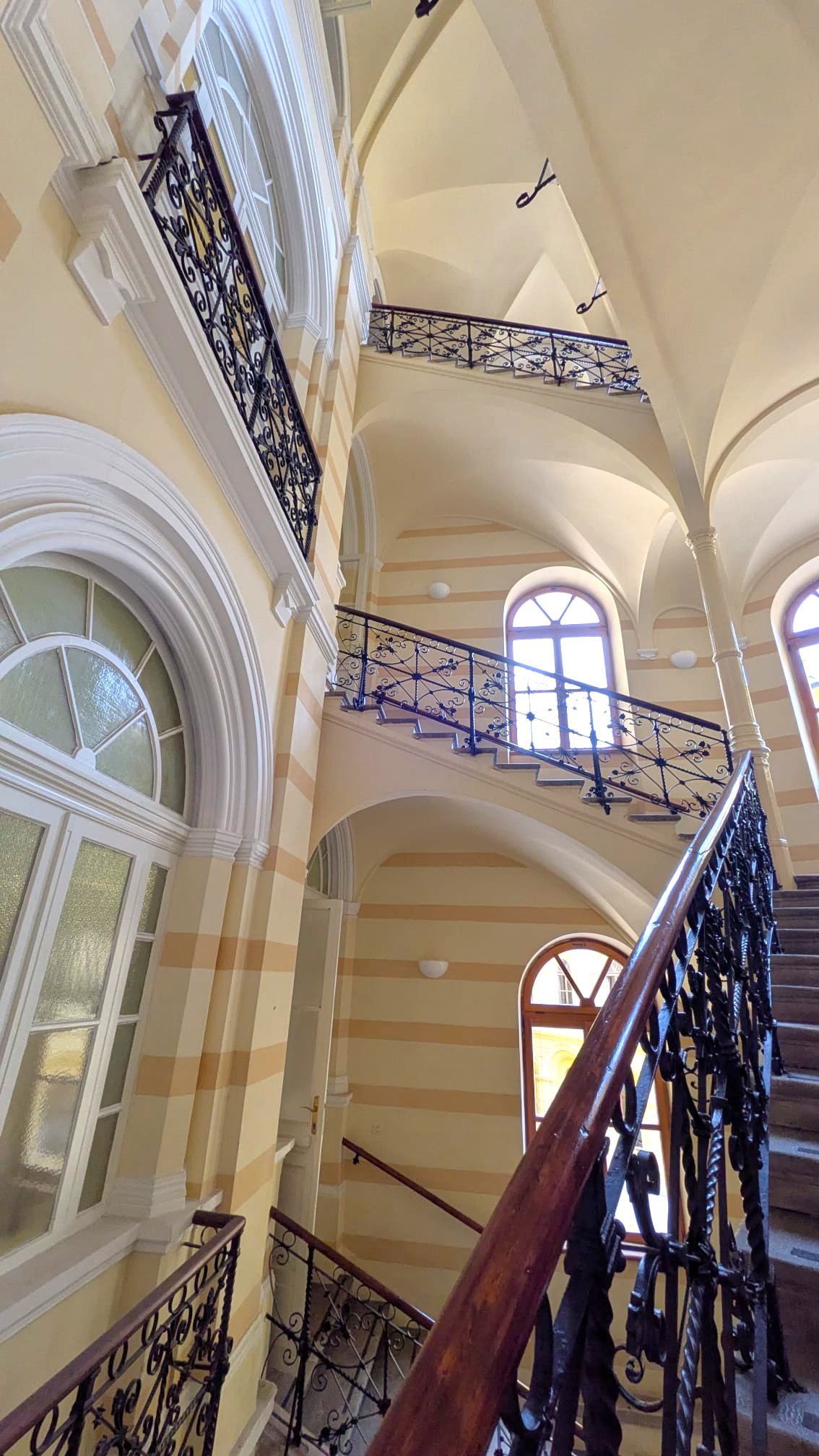 Interno di una scala ornata, con ringhiere in ferro battuto e pareti color crema e strisce. La luce naturale entra attraverso ampie finestre.