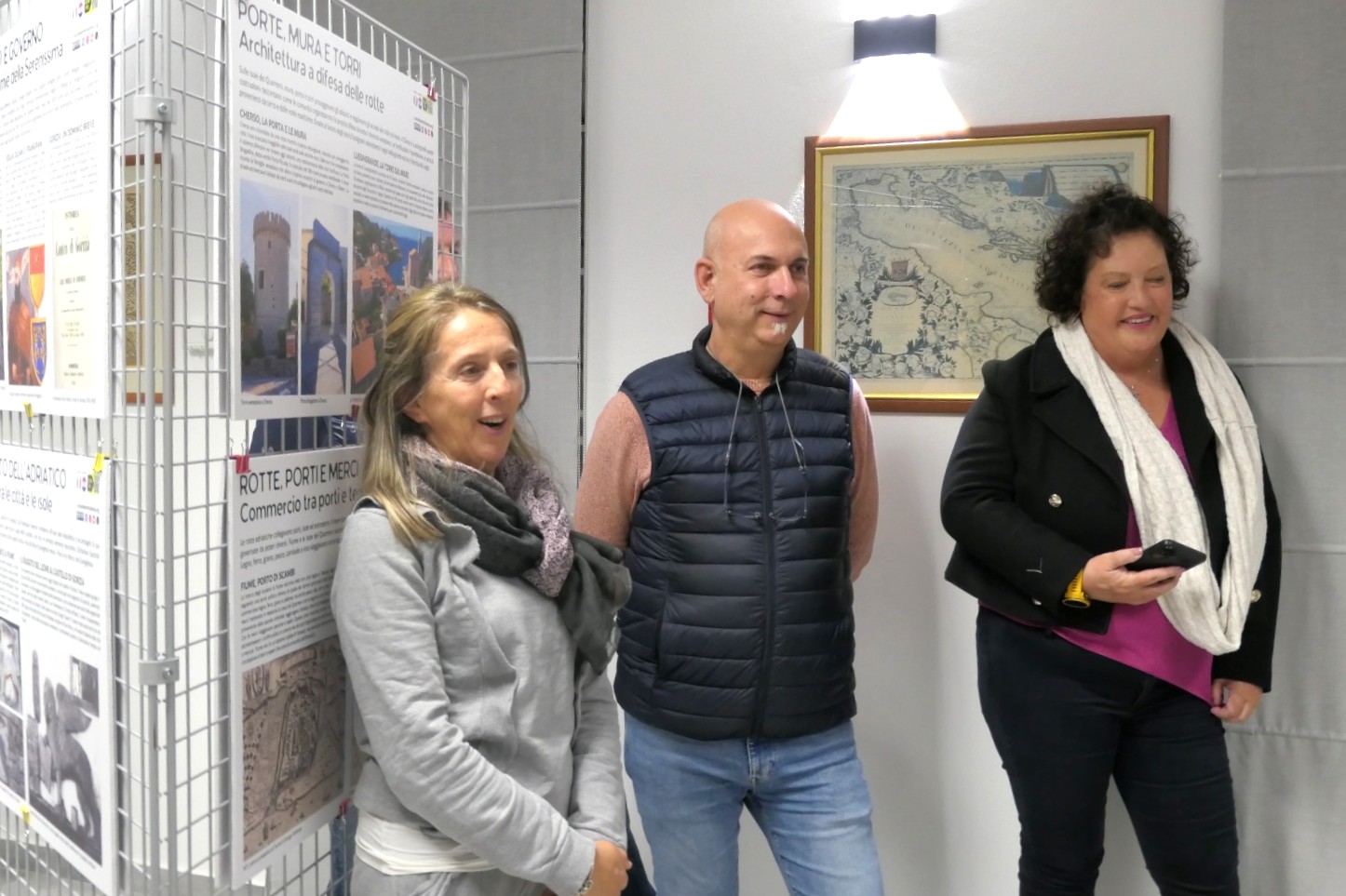 Tre persone sorridenti davanti a un'esposizione, con poster informativi sullo sfondo e un quadro appeso al muro.