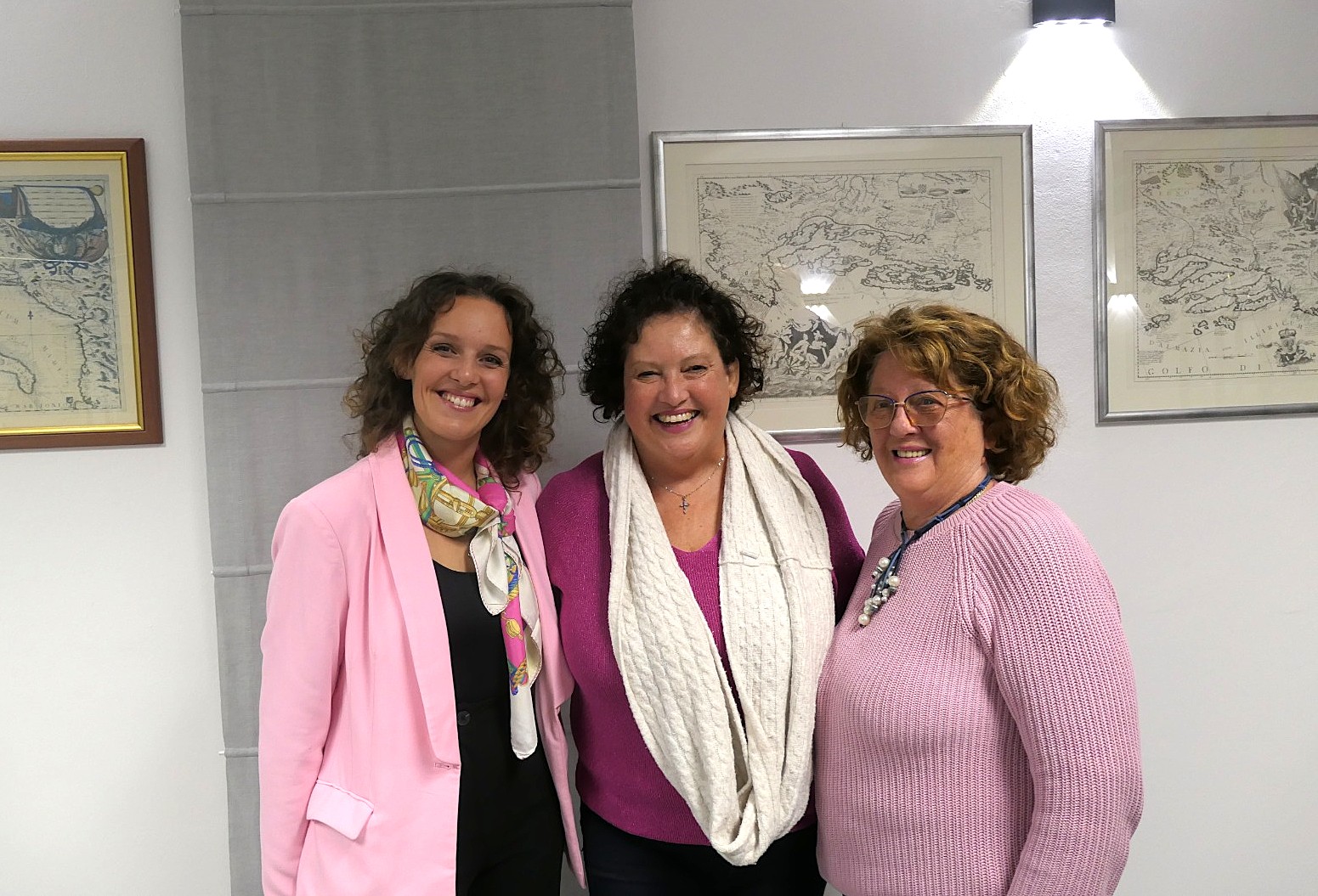 Tre donne sorridenti che posano insieme in un ambiente interno, vestite con abiti colorati e accessori, con mappe appese alle pareti.