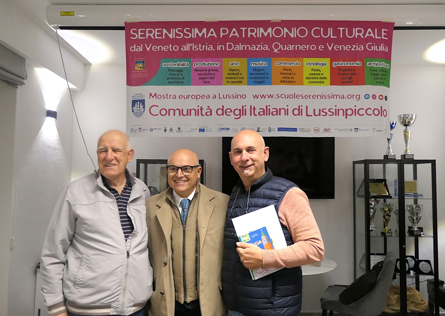 Tre uomini sorridenti in un ambiente interno, con un poster sopra di loro che promuove il patrimonio culturale della regione veneta e istriana.