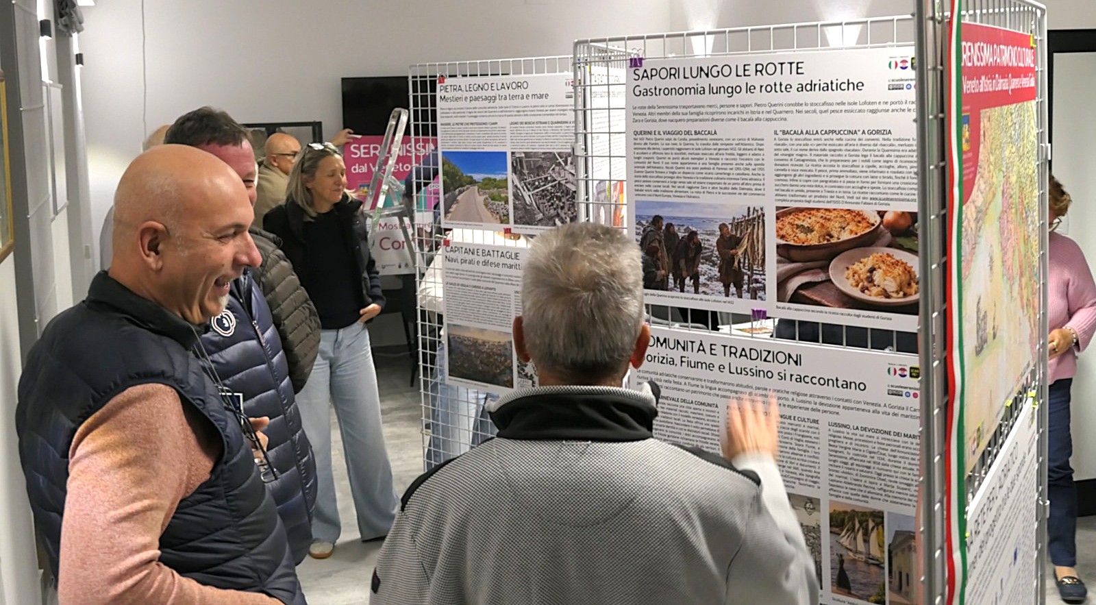 Persone che interagiscono di fronte a espositori con informazioni sulla gastronomia e tradizioni lungo le rotte adriatiche.