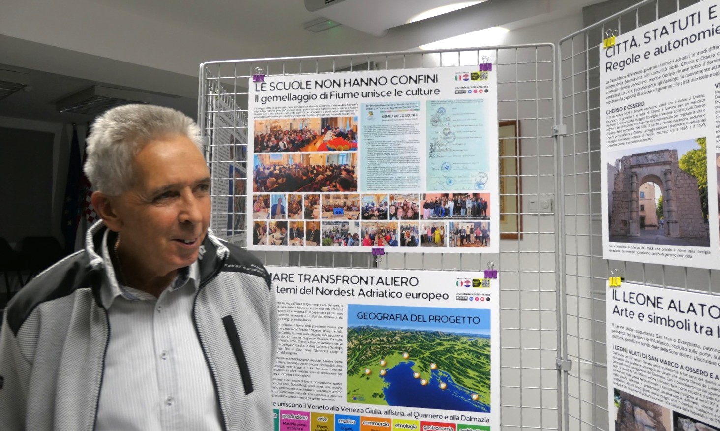 Un uomo sorride di fronte a un'esposizione di poster informativi su gemellaggi scolastici e cultura, in una sala luminosa.