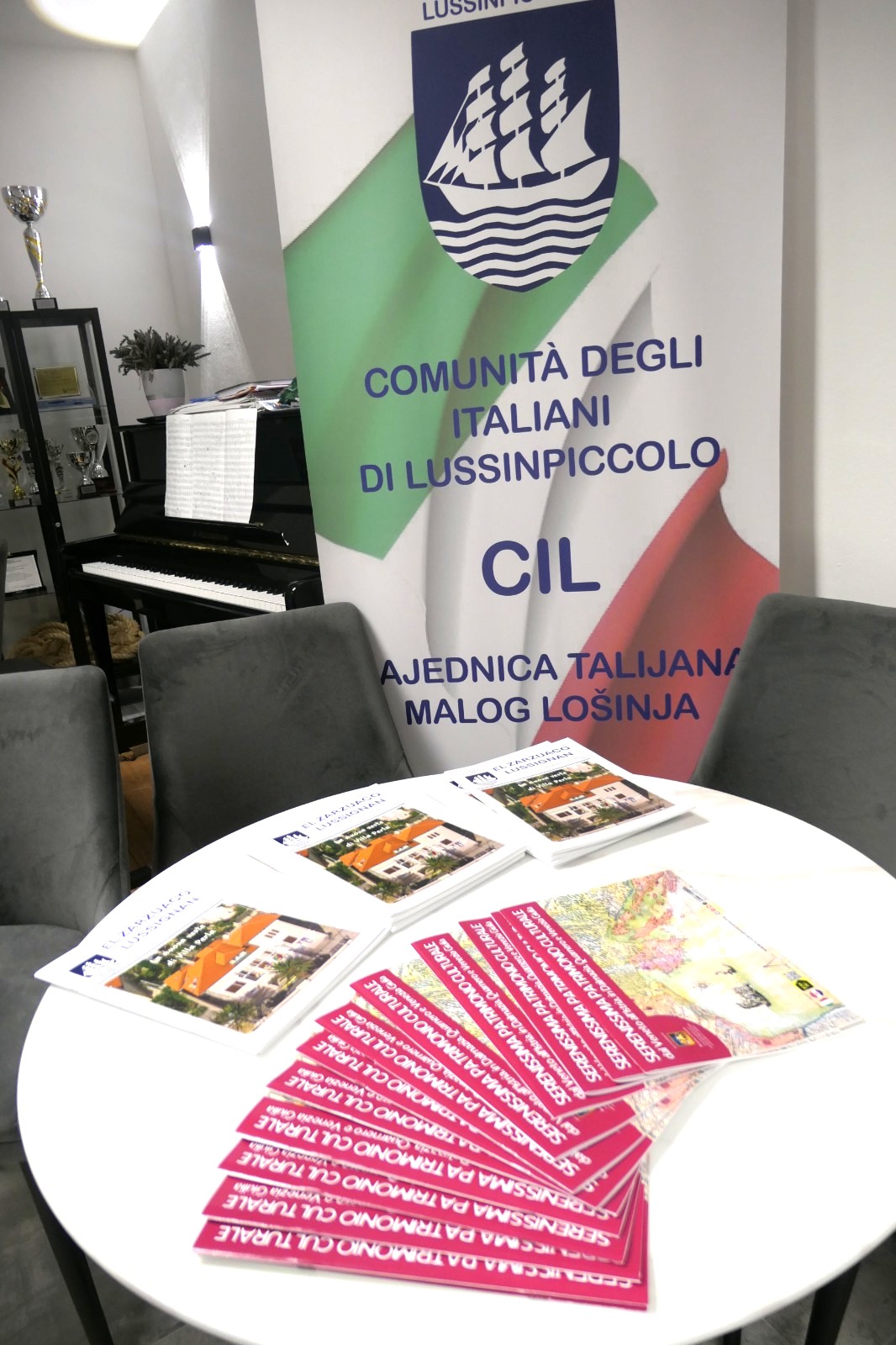 Striscione della Comunità degli Italiani di Lussinpiccolo con bandiera italiana sullo sfondo, con opuscoli su un tavolo bianco.