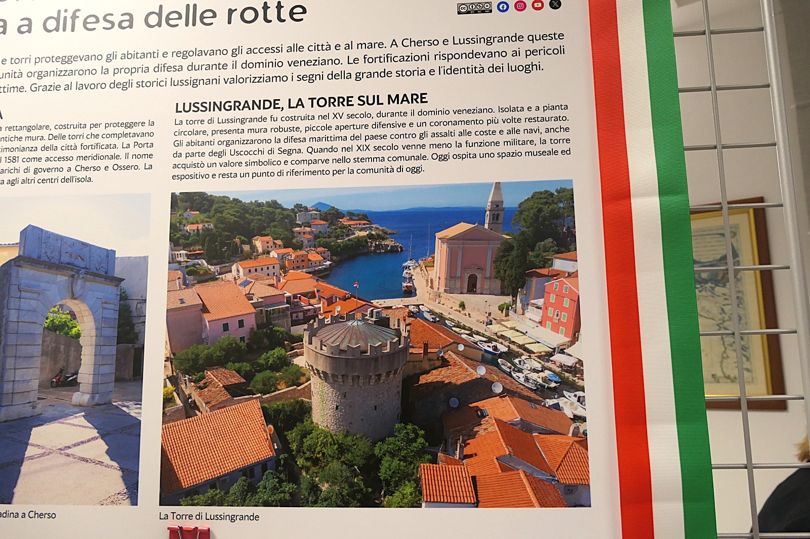 Immagine di un pannello informativo su fortificazioni storiche a Cherso e Lussingrande, con un focus sulla Torre di Lussingrande e il suo paesaggio circostante, visibile da un'angolazione aerea.