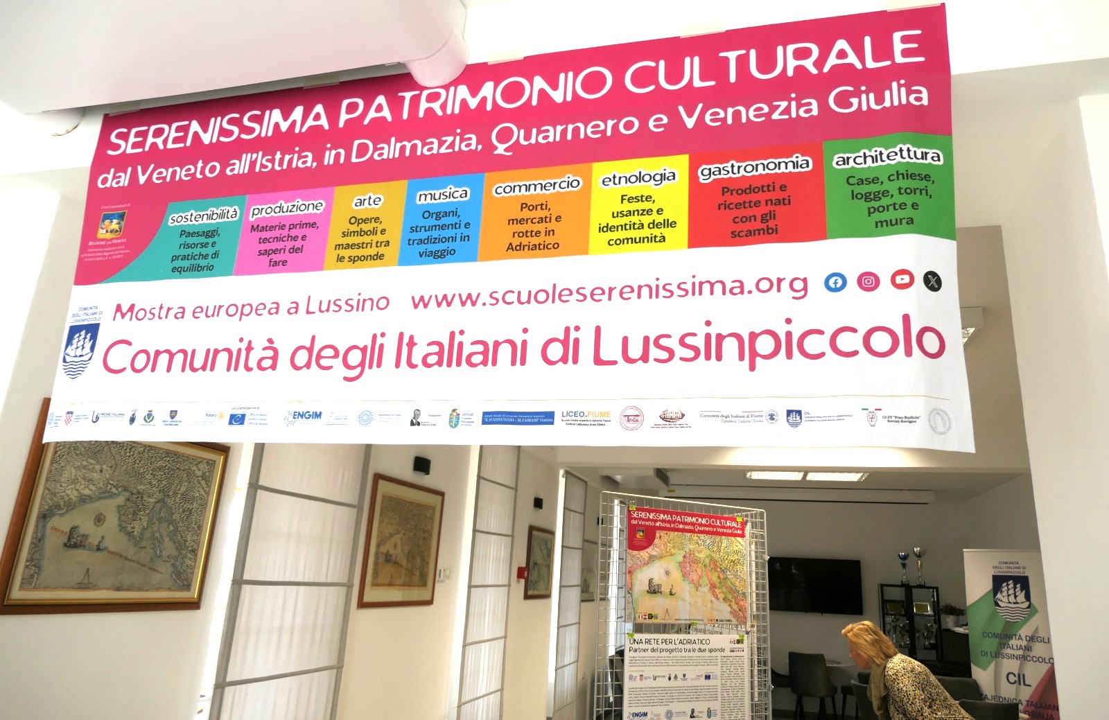 Striscione della mostra 'Serenissima Patrimonio Culturale' che espone temi di sostenibilità, arte, musica e gastronomia, presso un centro culturale.