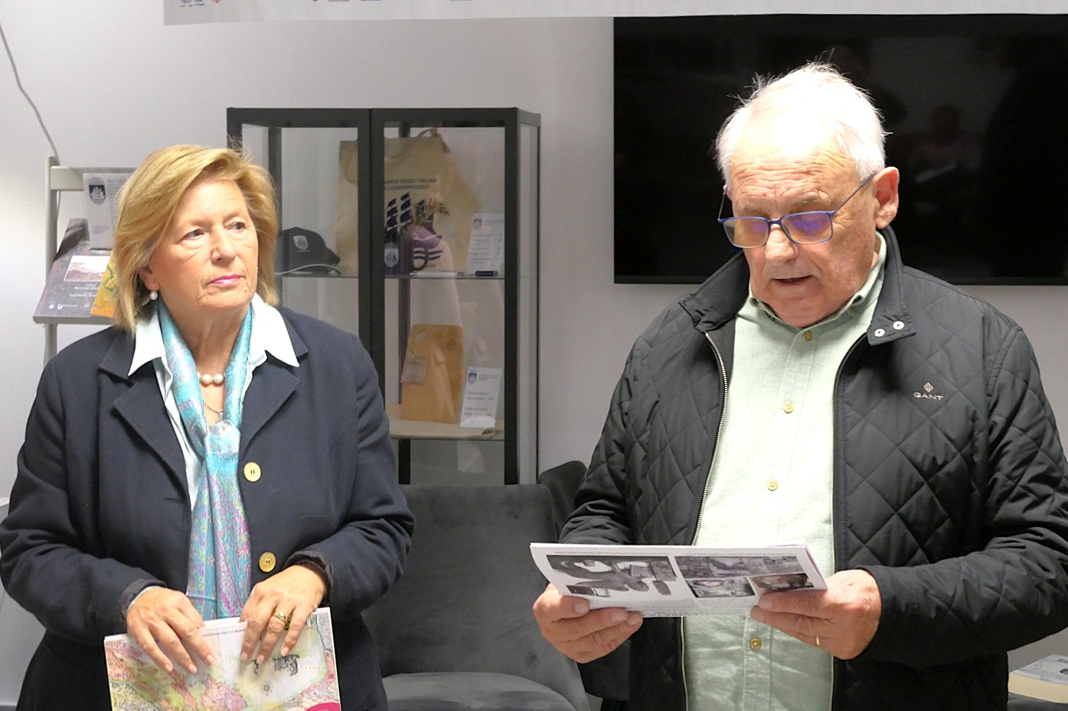 Due persone che partecipano a un evento, una donna in giacca scura e una sciarpa, e un uomo che legge un documento, con un espositore sullo sfondo.