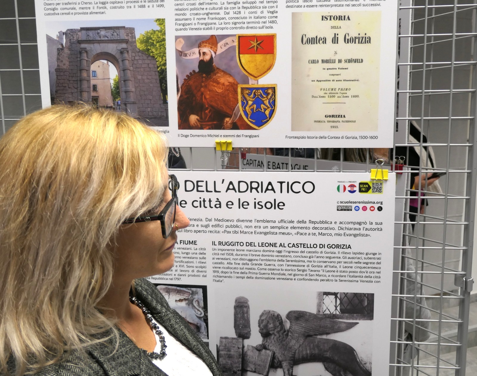 Donna con occhiali che osserva un pannello informativo sulla storia della Contea di Gorizia, con immagini e testi descrittivi su monumenti e personaggi storici.