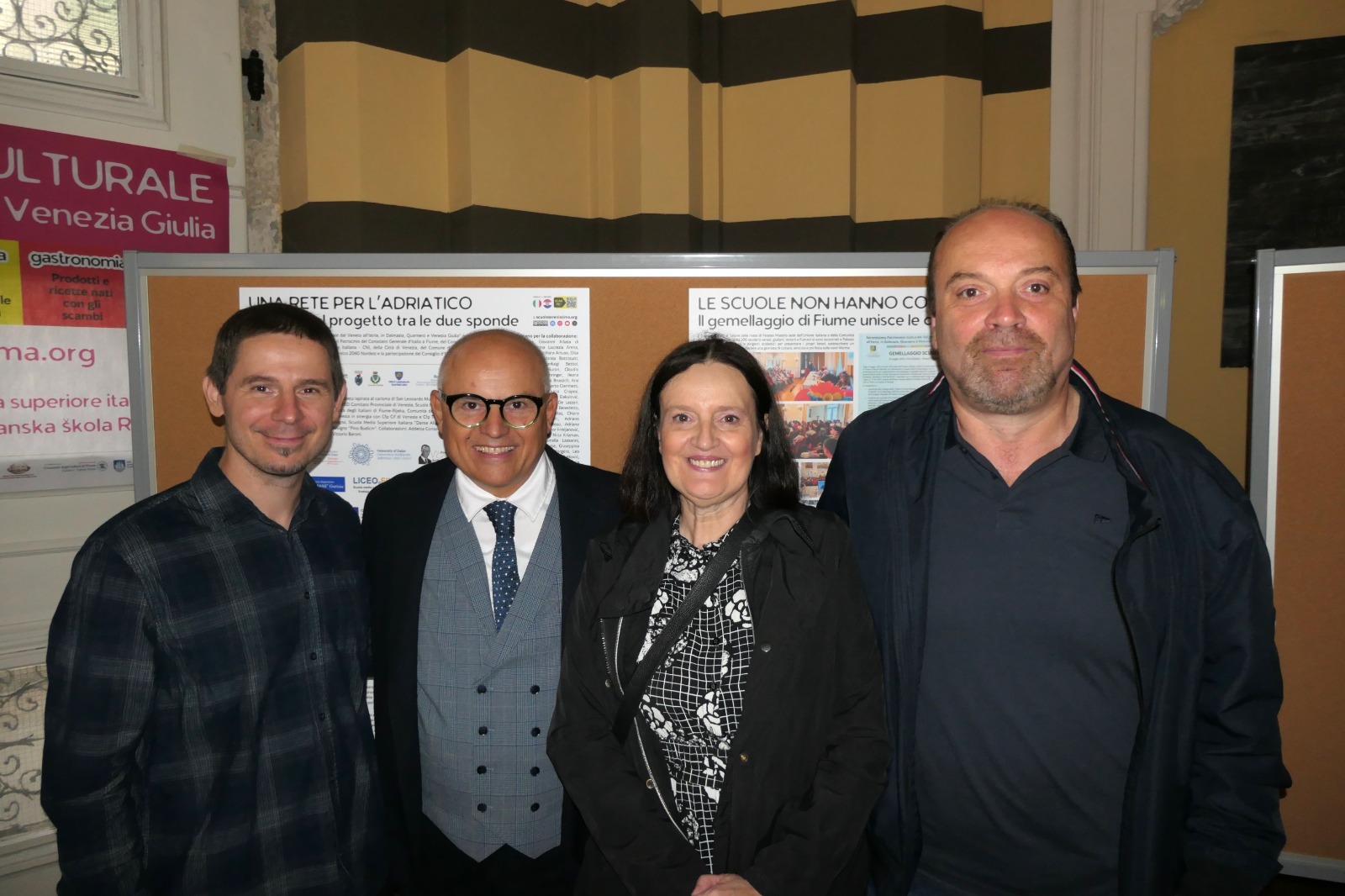 Quattro persone sorridenti posano per una foto davanti a un pannello informativo su un progetto culturale, in un ambiente interno con pareti a strisce.