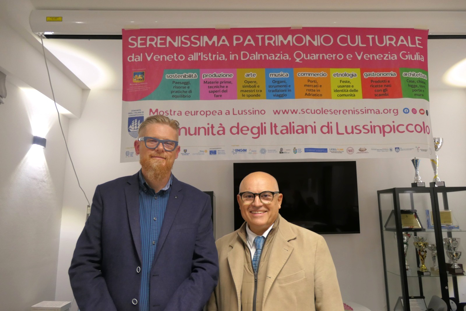 Due uomini sorridenti in piedi davanti a un banner che promuove una mostra culturale a Lussinpiccolo, evidenziando elementi di patrimonio e tradizioni delle comunità italiane dell'Istria, della Dalmazia, del Quarnaro e della Venezia Giulia.