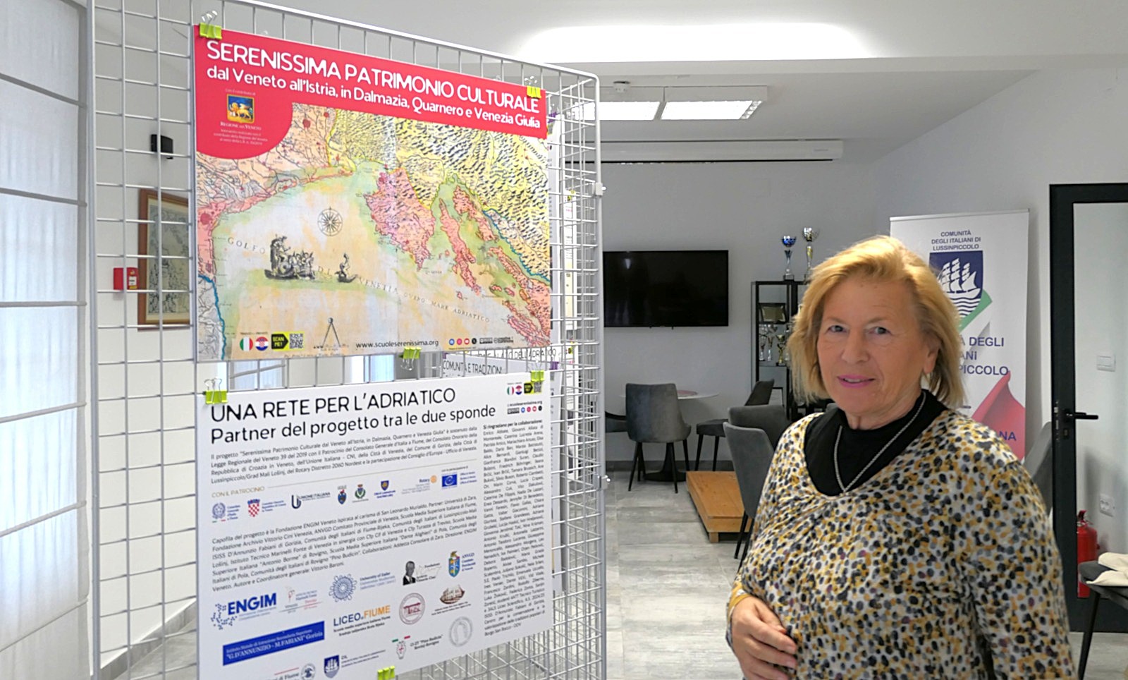 Una donna sorridente di fronte a un poster informativo sulla Serenissima, con una mappa storica della regione adriatica visibile sullo sfondo.