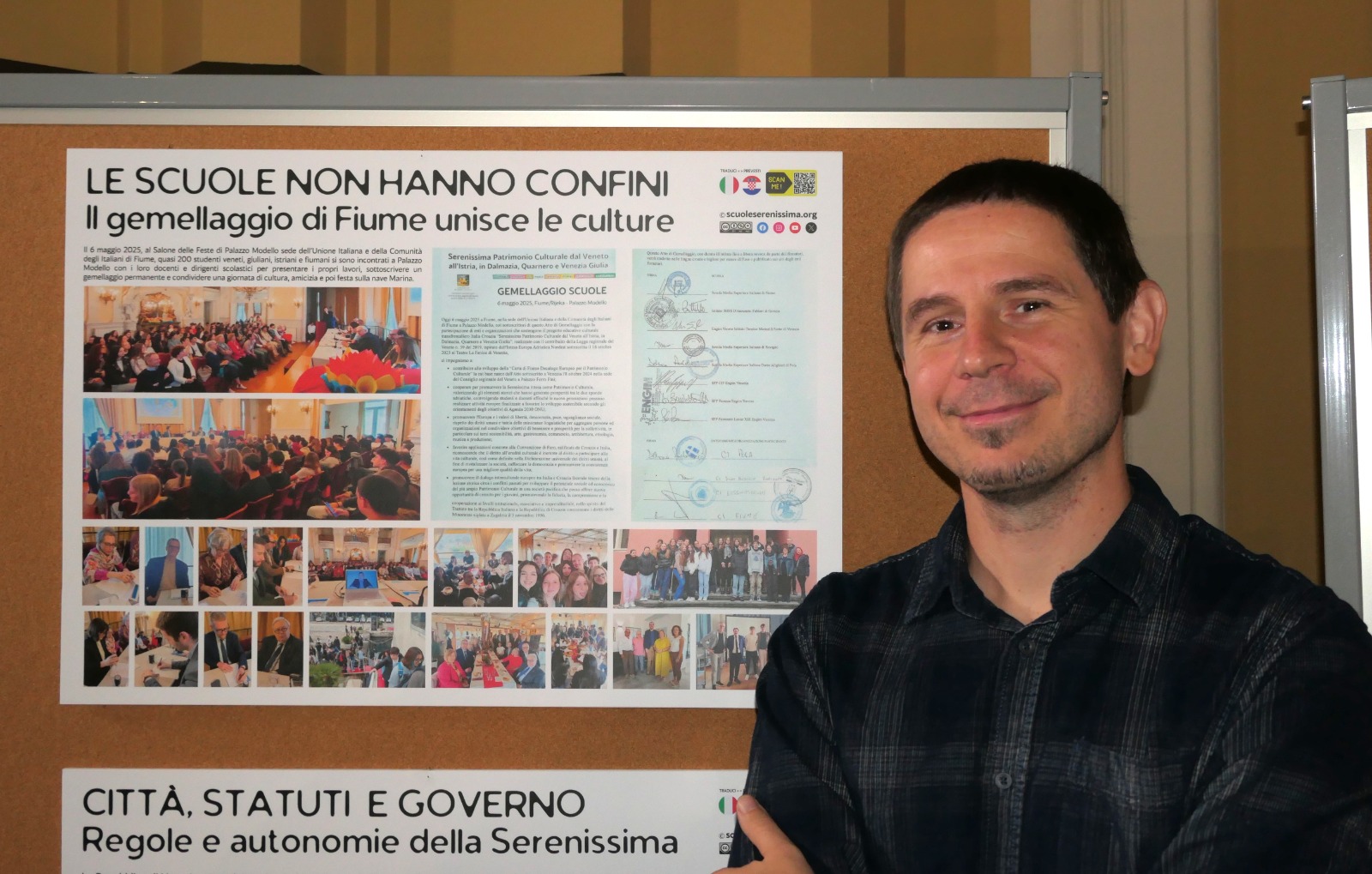 Un uomo sorride accanto a un poster che promuove il gemellaggio di Fiume, evidenziando l'importanza della cultura e dell'istruzione senza confini.