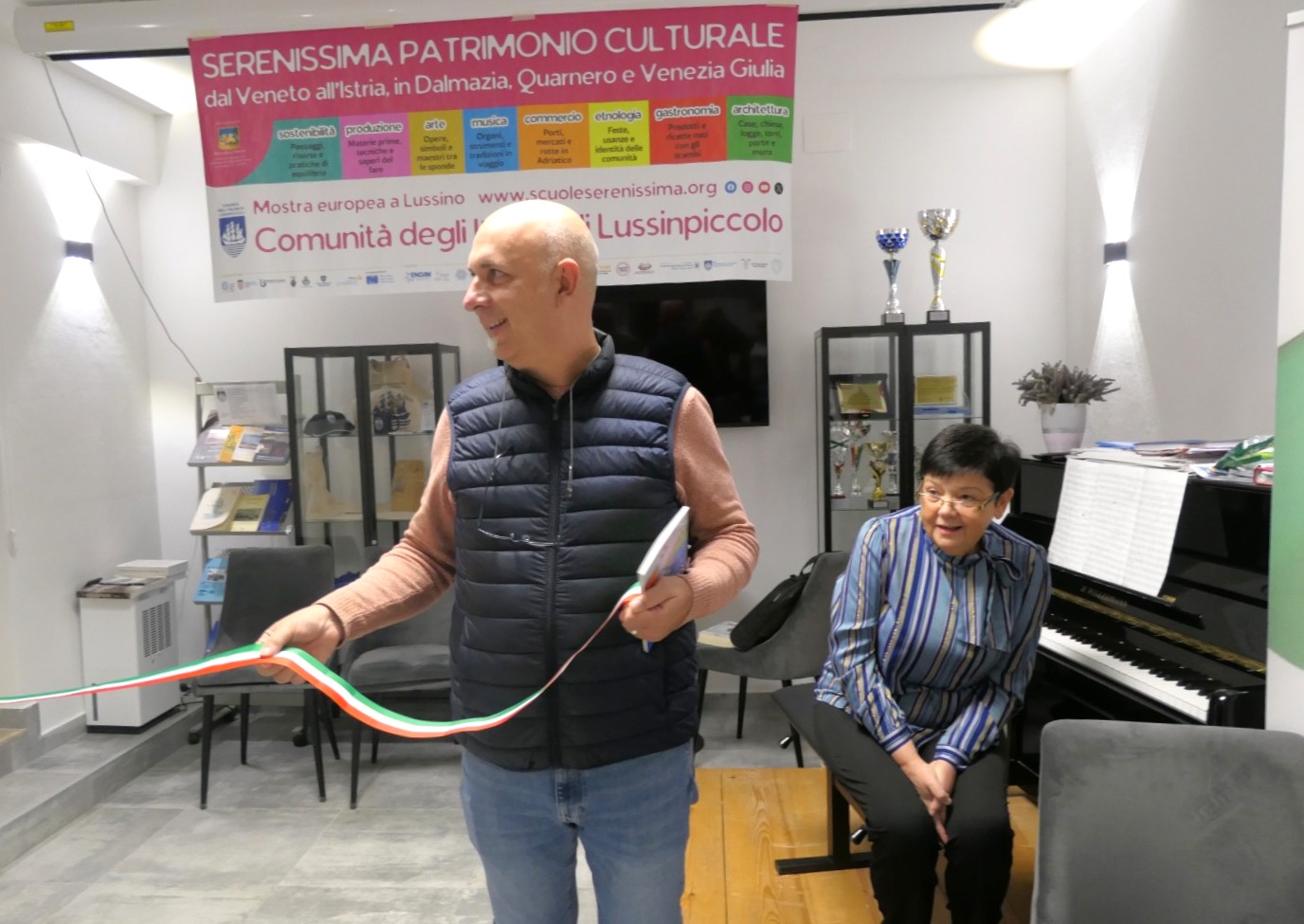 Due persone in una sala, una tiene un nastro tricolore, l'altra è seduta vicino a un pianoforte. Sullo sfondo, un poster che promuove il patrimonio culturale dell'area.