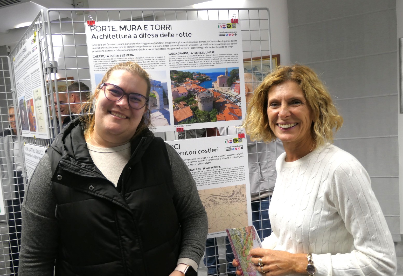 Due donne sorridenti davanti a un'esposizione di pannelli informativi su architettura costiera, tra cui riferimenti a porte, mura e torri.